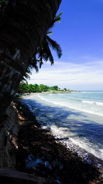 Bright skies show the shoreline of Mui Ne in Lam Dong. Waves roll onto the beach as palm trees provide shade. People enjoy the sunny day by the water and explore the area.