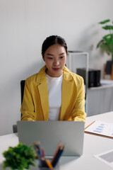 Asian businesswoman wearing a yellow blazer, working diligently on a laptop in a modern office,...