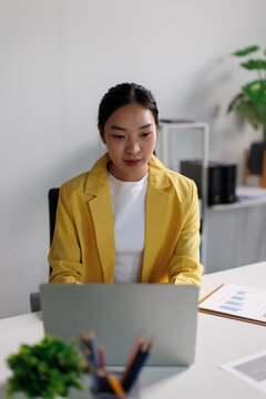Asian businesswoman wearing a yellow blazer, working diligently on a laptop in a modern office, embodying dedication, efficiency, and professional success in her career