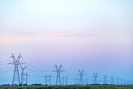 Row of high voltage power transmission towers and power lines stretching across rural field at twilight in USA