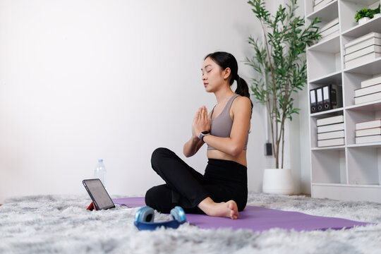 Young asian woman practicing yoga and meditation at home, finding inner peace and mindful wellness, following an online fitness class on a digital tablet for spiritual calm