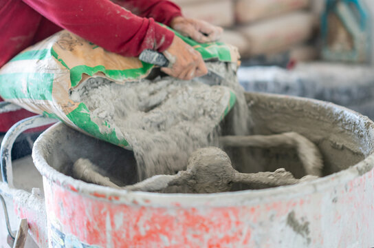 Worker hands emptying a bag of dry mortar mix into an active cement mixer drum, preparing concrete batch for building renovation work.