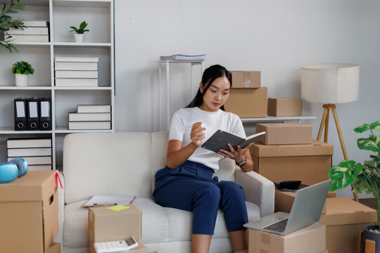 Young asian woman working from her home office, sitting on a sofa and reviewing information in a notebook, surrounded by cardboard boxes for shipping online orders