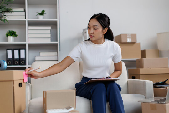 Asian female entrepreneur preparing e commerce parcel for shipping from home office, checking inventory, managing online orders, small business owner working remotely