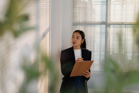 Asian businesswoman in a modern office holding a clipboard and pen, gazing thoughtfully out the window while planning projects and focusing on career goals and strategy