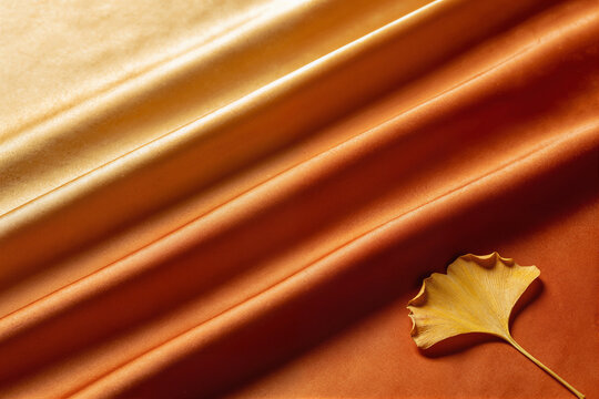 Smooth orange satin fabric with flowing folds and a single ginkgo leaf, elegant autumn background