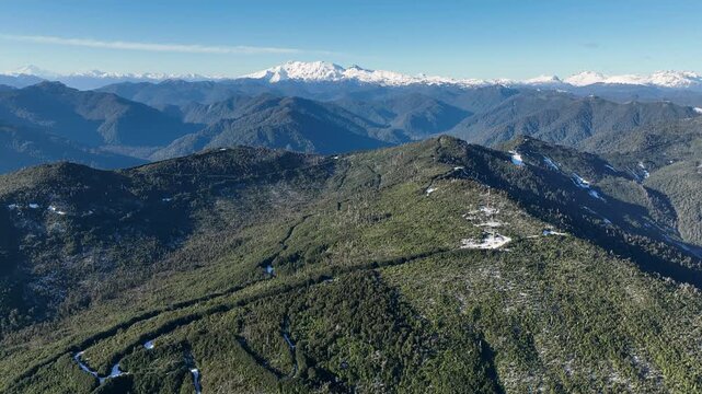 A drone pans slowly across a forested Andes ridge in southern Chile, revealing deep valleys and patchy snow, with the majestic Volc&aacute;n Yate rising on the horizon.