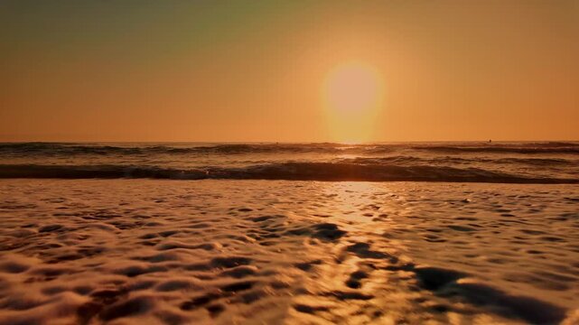 Gentle ocean waves cascade over wet sand under a vibrant golden sunset, reflecting warm light across the water and shoreline.