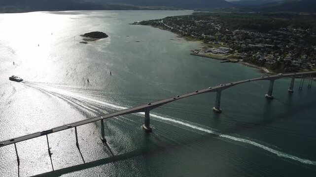 Aerial drone footage looks down over the Gisund Bridge in Norway as a ferry passes beneath it, leaving a long wake, while cars cross above toward Senja island