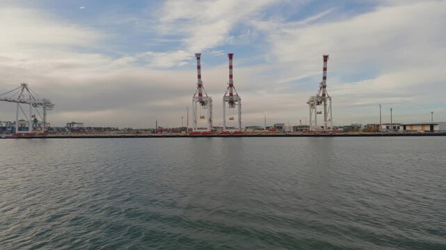 Large cranes stand tall against a cloudy sky. Water ripples gently beneath the industrial skyline. Dockside activity continues quietly, unbothered by passing ships. Ideal for maritime or trade visuals
