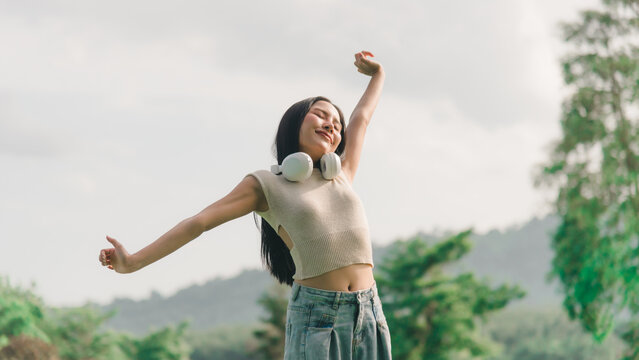 Happy woman stretching in nature, raising arms outdoors, relaxing muscles while wearing headphones, enjoying serene moment of freedom, breathing fresh air and feeling peaceful body wellness