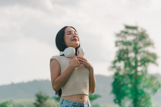 Woman breath healing careful praying inner peace while smiling outdoors enjoying calm natural environment with white headphones resting around neck during summer day relaxation routine