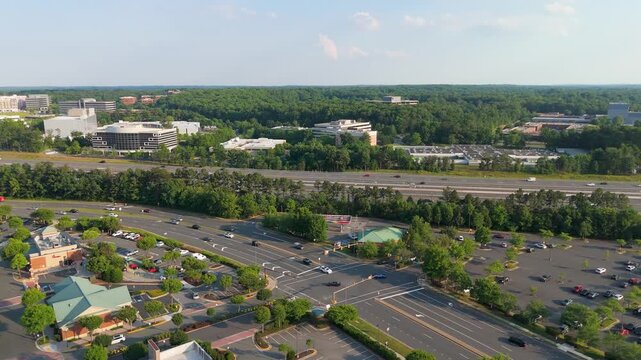 Aerial drone view of busy suburban intersection traffic, retail parking lots, and commercial office parks surrounded by green trees in Reston and Herndon, Northern Virginia on a sunny day.