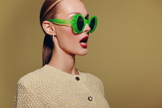 Stylish woman in green sunglasses and beige textured jacket looks surprised with open mouth on plain background. Concept of fashion, retro style, and expressive emotions in studio portrait.