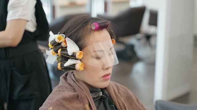 Hairdresser wearing gloves places rubber basin near woman's shoulder during perm neutralizing process with rollers