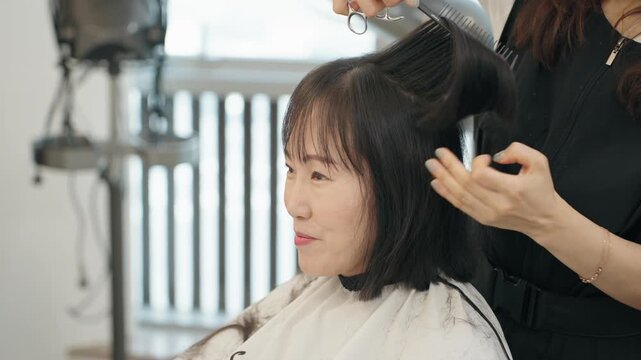 Hairstylist lifts and combs woman's dark bob hair while trimming top section with scissors during haircut