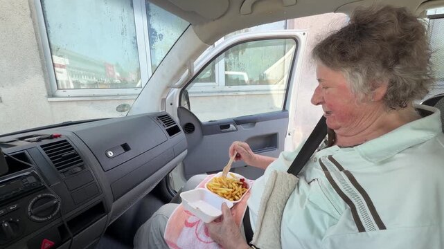 Elderly woman sits inside a van with the side door open while eating a portion of French fries with ketchup and mayonnaise using a small wooden fork. She pauses while eating. Static shot