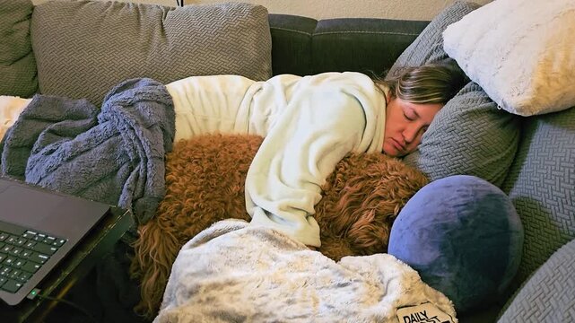 Relaxed woman naps beside her curly brown dog on a comfortable sofa, wrapped in blankets with plush pillows and a nearby laptop in a quiet home interior for cozy lifestyle footage.