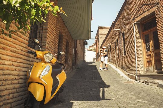 Young couple kissing beside yellow motor scooter. Romantic cobblestone alleyway, old Tbilisi architecture. Summer vacation, Georgia travel destination, authentic city lifestyle