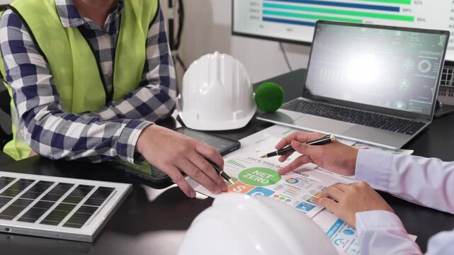 Two engineers in safety vests discussing ESG green energy strategy and solar cell technology at desk with tablet and house model.