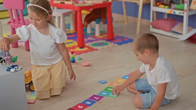 Girl and brother sit and kneel on a wooden floor arranging numbered cards and toy truck beside a shelf and puzzle mat in building; playful bonding.