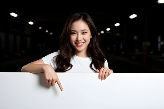 Cheerful cute girl is standing behind the white blank banner and pointing down at a copyspace