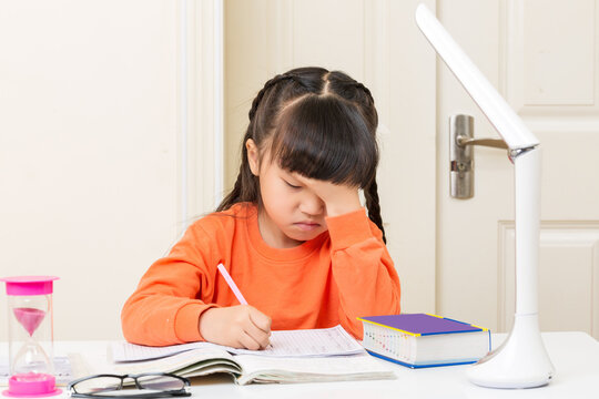 Stressed little East Asian girl holding her forehead while doing difficult homework at her desk.