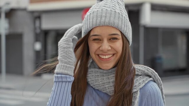 Woman smiling with gloved hand touching face, wearing beanie, scarf and sweater on a busy city street near a storefront and parked car; cozy joy.