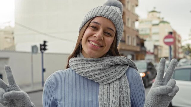 Woman wearing gray knit scarf, pompom beanie and gloves holds up peace sign on a city street while smiling broadly; cozy joy.