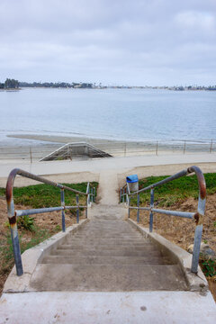 Concrete stairs with metal handrails leading down to a scenic coastal Mission Bay pathway under a cloudy sky, San Diego, CA