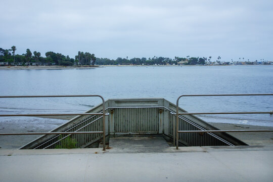 A concrete pedestrian ramp structure with metal railings leads down from a beachfront walkway toward a calm Mission Bay under cloudy skies, San Diego, CA
