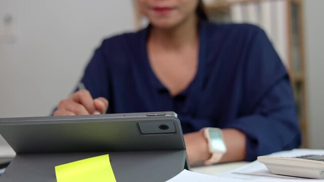 Asian female worker tapping tablet screen to open business presentation file while sitting at white office table near sticky memo note in bright corporate setting space room