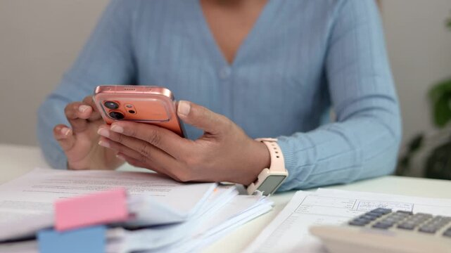 Asian female employee browsing application on mobile phone while sitting at desk in bright modern office searching information and coordinating with team during busy workday