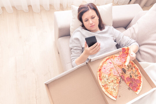 Depressed woman eating pizza and holding phone laying on sofa at home, warm yellow tone