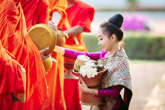Young Laotian woman in traditional silk dress giving morning alms sticky rice to Buddhist monks in Luang Prabang, Laos travel banner concept.