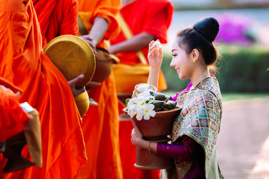 Young Laotian woman in traditional silk dress giving morning alms sticky rice to Buddhist monks in Luang Prabang, Laos travel banner concept.