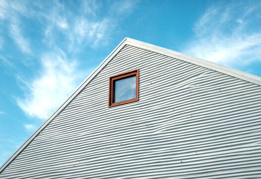 Modern metal roof with small attic window under clear blue sky with soft white clouds