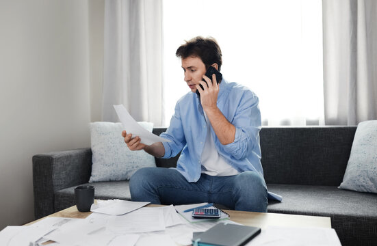 Stressed man on the phone looks shocked at utility bills due to rising energy prices and inflation. He struggles with household expenses, overdue bills, and financial problems at home.