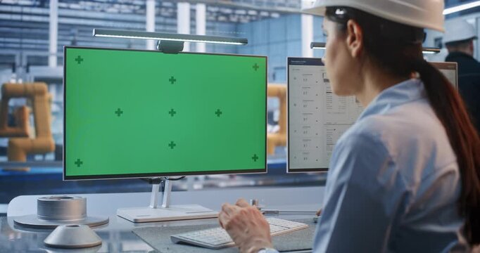 Inside Modern Smart Factory, Young Female Engineer in Hard Hat Operates PC with Chroma Key Green Screen Monitor. Ideal for Compositing Customized Industrial Software and Telemetry. AI Manufacturing.