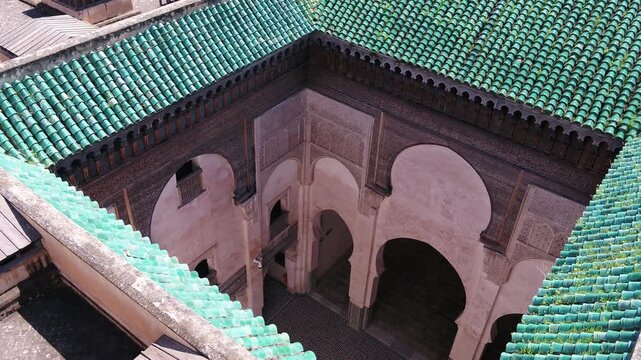 Close elevated drone perspective capturing intricate carved cedar corbels, horseshoe arches, and green glazed tile roofs of a historic madrasa in Fez, Morocco.