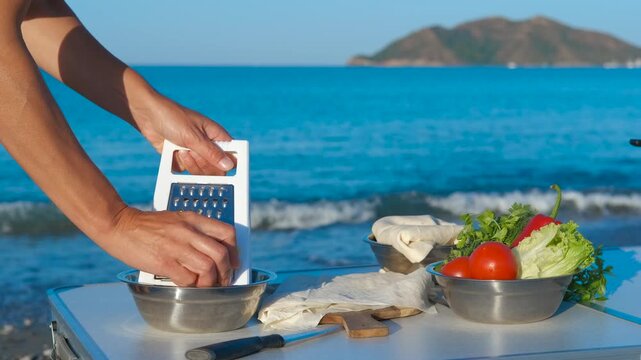 Chef grating cheese on the beach for delicious meal. On a sunny beach, a chef expertly grates cheese over a bowl, preparing a meal with fresh vegetables and flatbread, the tranquil sea a backdrop