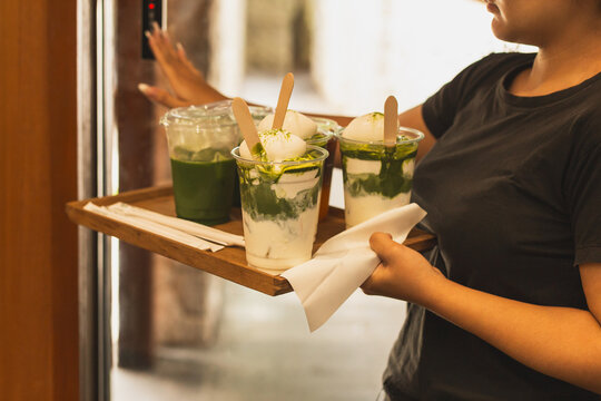 Waitress carrying a wooden tray with iced matcha drinks while pushing door button in a cafe.
