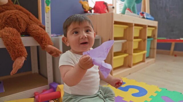 Toddler boy holding purple paper and small toy on foam number mat in playroom building; curiosity play learning.