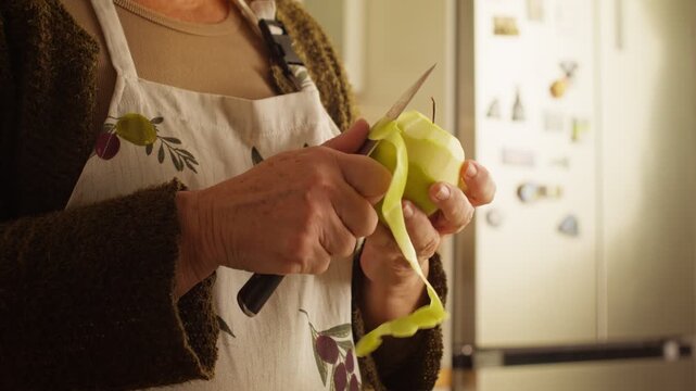 Hands peel a green apple with a kitchen knife under warm indoor lighting