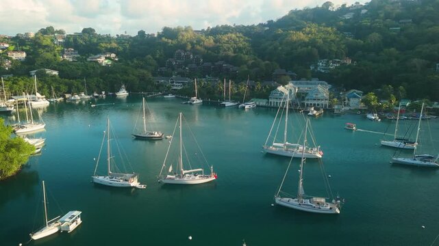 Drone performs close parallax shot around anchored sailboats in Marigot Bay marina with hillside resort and lush greenery in background