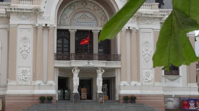 Close-up view of Saigon Opera House entrance with caryatid statues framed by green leaves, Ho Chi Minh City, Vietnam 