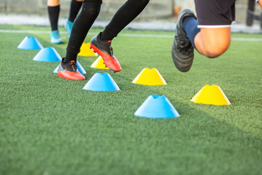 Selective focus to legs of young soccer players practicing agility drills, running past blue and yellow marker cones on a green artificial turf field during football training session.