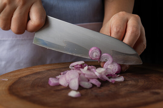 Close up of sharp chef knife blade slicing fresh purple shallots on wooden board.