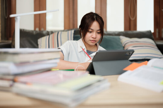Asian teenage girl concentrates on online class, remote learning at home using tablet, writing and reading textbooks, self-study for back to school, Homeschooling, university education lifestyle.