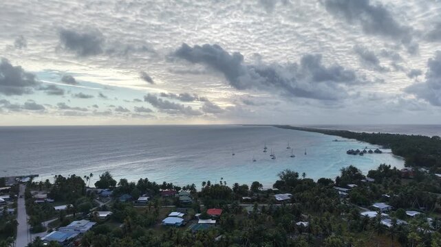 Aerial descending over a turquoise lagoon, showcasing anchored sailboats, coastal village houses, and the Kia Ora Resort in Rangiroa Atoll